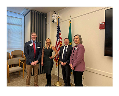 Four people posing indoors next to an American flag and Washington State flag during a legislative meeting at the Capitol